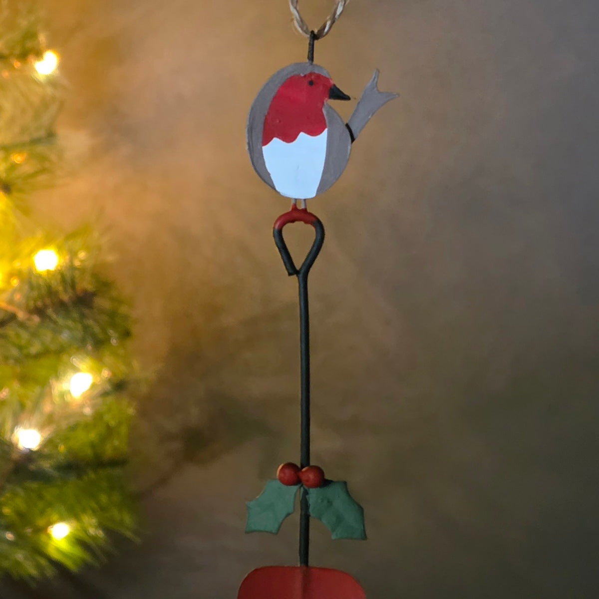 A hanging Christmas ornament shaped like a robin perched on a metal spade, with holly leaves and berries near the bottom, shown against a softly blurred holiday background with warm lights.