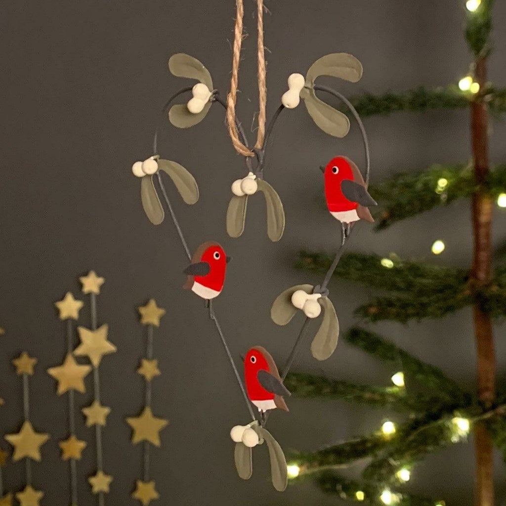 A metal hanging Christmas heart shaped garland decoration with mistletoe and robin decorations, shown against a dark indoor background with blurred holiday lights and tree branches.