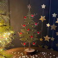 A decorative metal Christmas tree ornament in a green tiered silhouette on a wooden base, small gold ball accents, red robins displayed on a tabletop with holiday decor in the background.