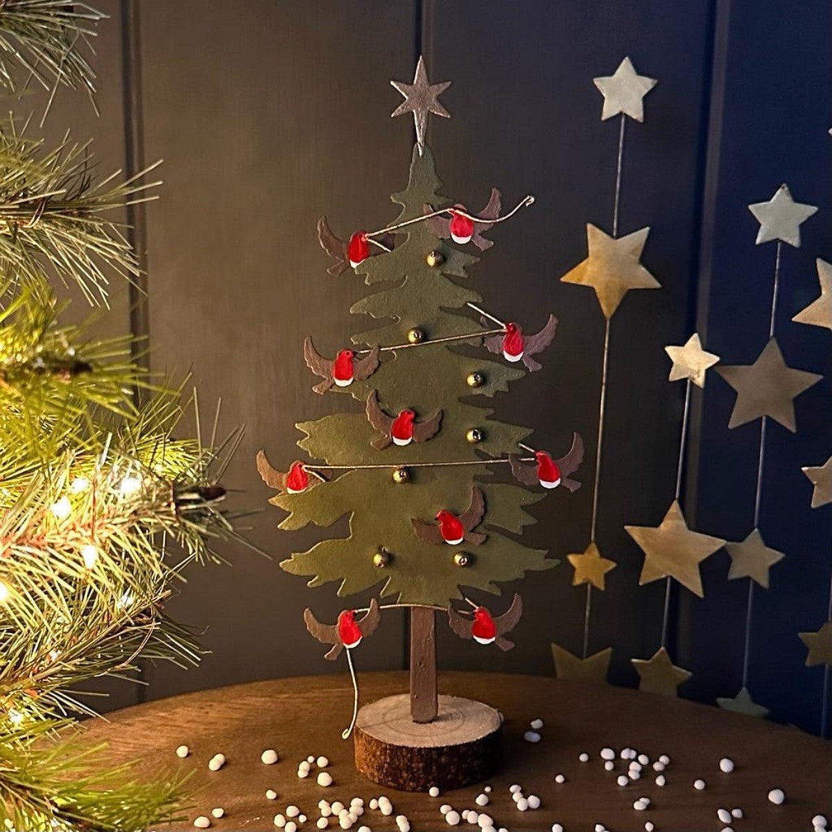 A decorative metal Christmas tree ornament in a green tiered silhouette on a wooden base, small gold ball accents, red robins displayed on a tabletop with holiday decor in the background.