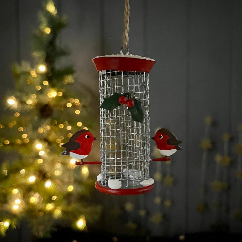 A metal bird feeder in red and silver with two robins perched either side with a holly leaf and berries motif decoration a the front 