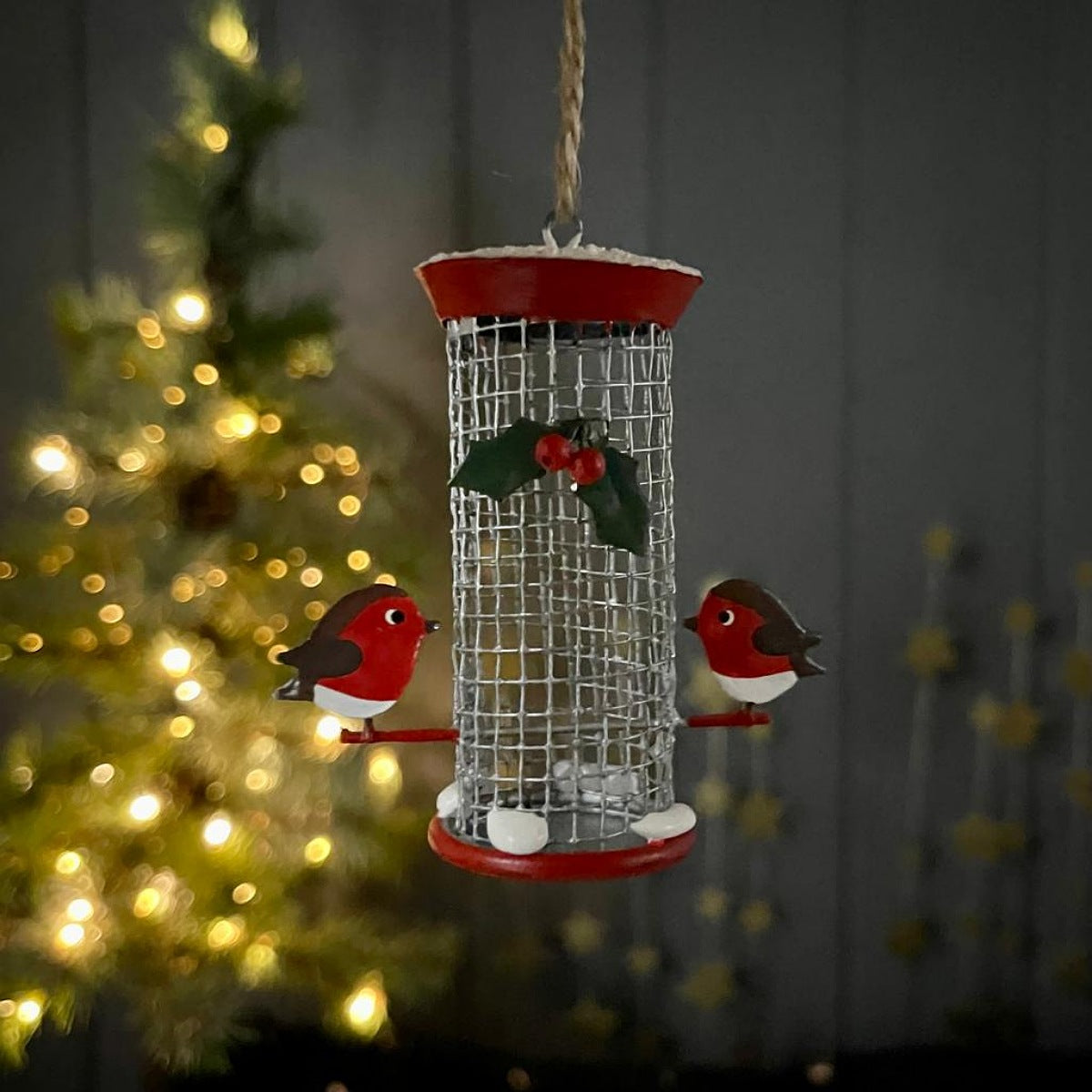 A metal bird feeder in red and silver with two robins perched either side with a holly leaf and berries motif decoration a the front 