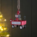 A Christmas metal hanging decoration shaped like a red wagon filled with wrapped presents, with a small bird figure on the side and a hanging twine loop at the top, shown against a dark panelled background with blurred warm lights on the left.