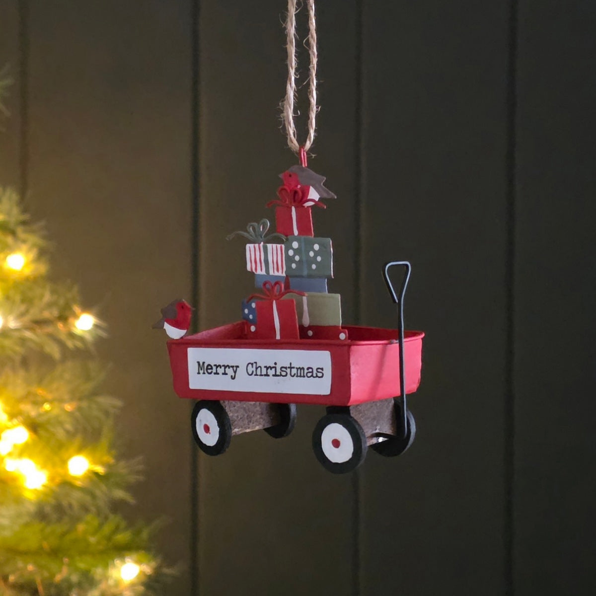 A Christmas metal hanging decoration shaped like a red wagon filled with wrapped presents, with a small bird figure on the side and a hanging twine loop at the top, shown against a dark panelled background with blurred warm lights on the left.