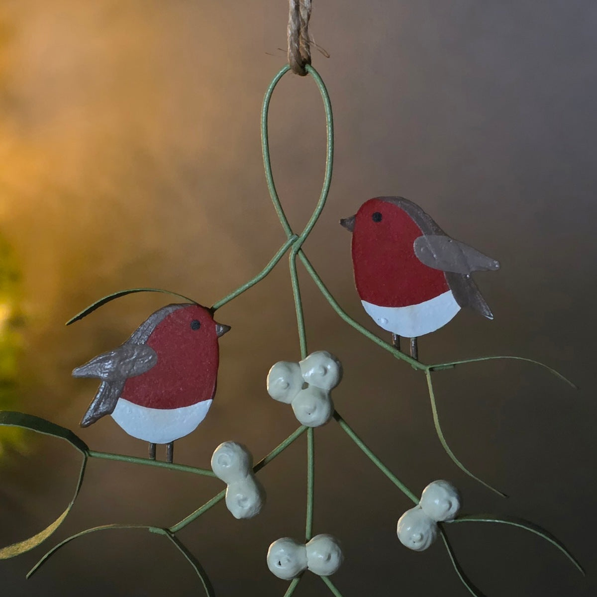 A hanging decorative ornament shaped like two small birds perched on green vine branches with white berry clusters, photographed against a softly blurred warm-toned background.