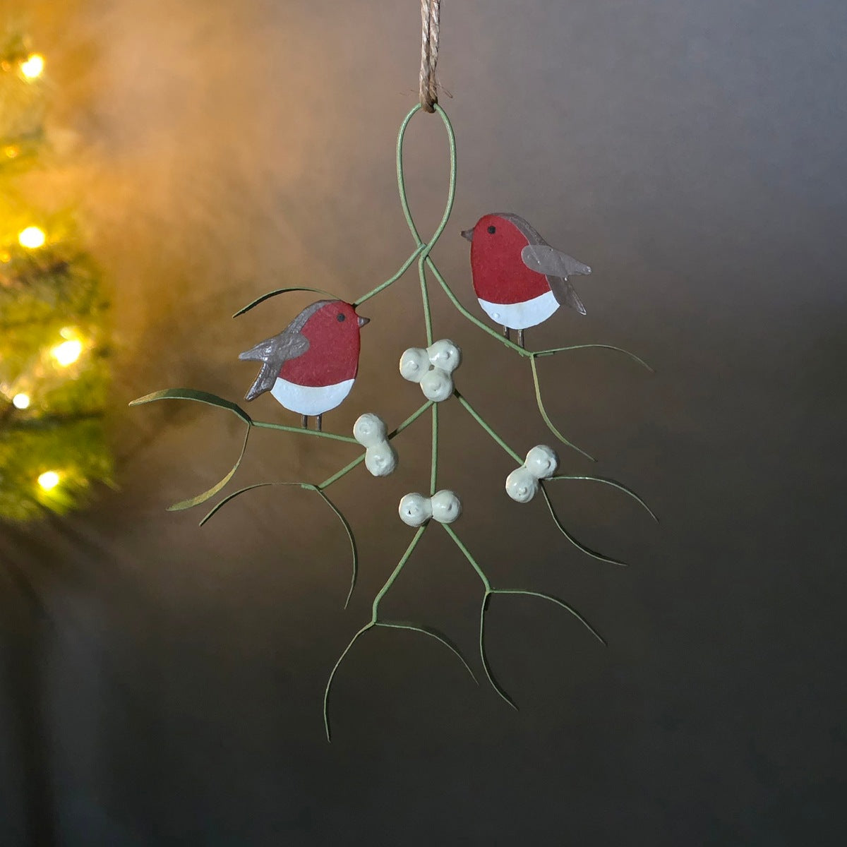 A hanging Christmas decoration featuring small red birds with gray wings sitting on a  mistletoe branch with white berries, shown against a dark indoor background with warm lights on the left.