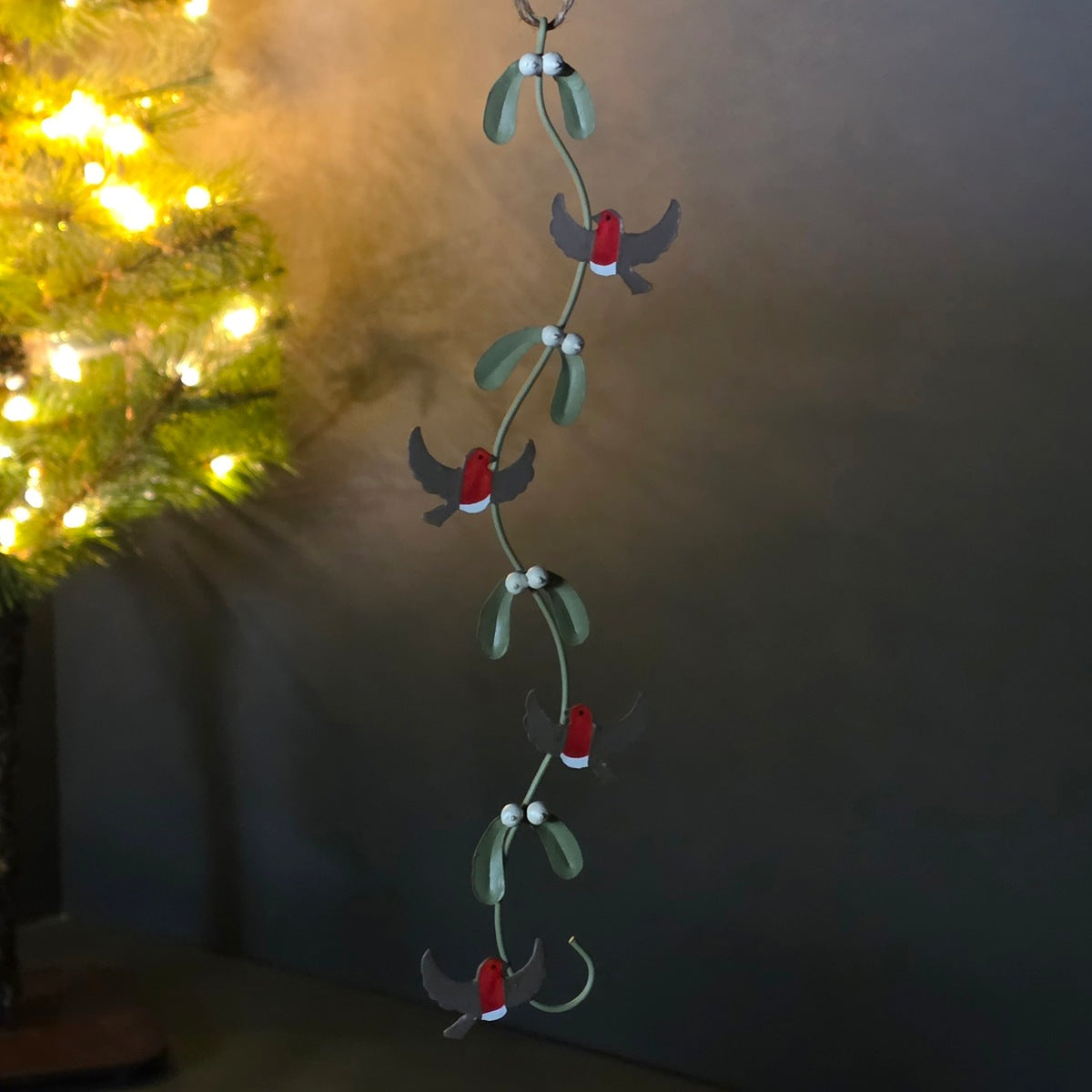 A hanging Christmas garland featuring small red birds with gray wings and green mistletoe leaves with white berries, shown against a dark indoor background with warm lights on the left.