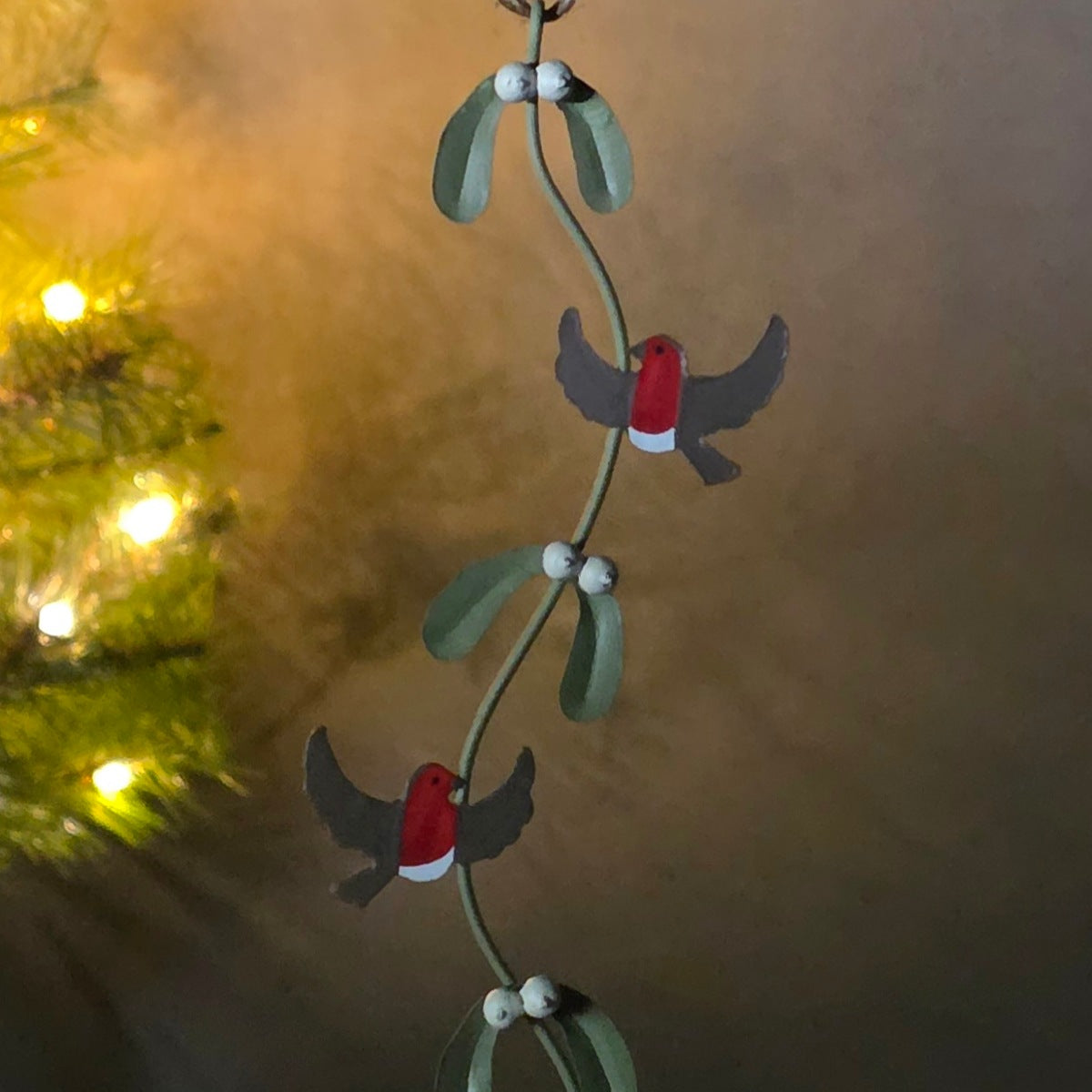 A hanging Christmas ornament featuring three small birds with red bodies, gray wings, and white underbellies perched on green mistletoe branches with white berries, shown against a warm blurred background with holiday lights.