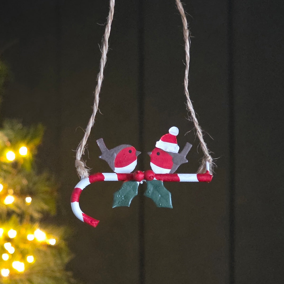 A hanging metal Christmas ornament featuring two small red and white robin birds perched on a candy cane, with green holly leaves and twine suspension, photographed against a dark background with blurred warm lights.