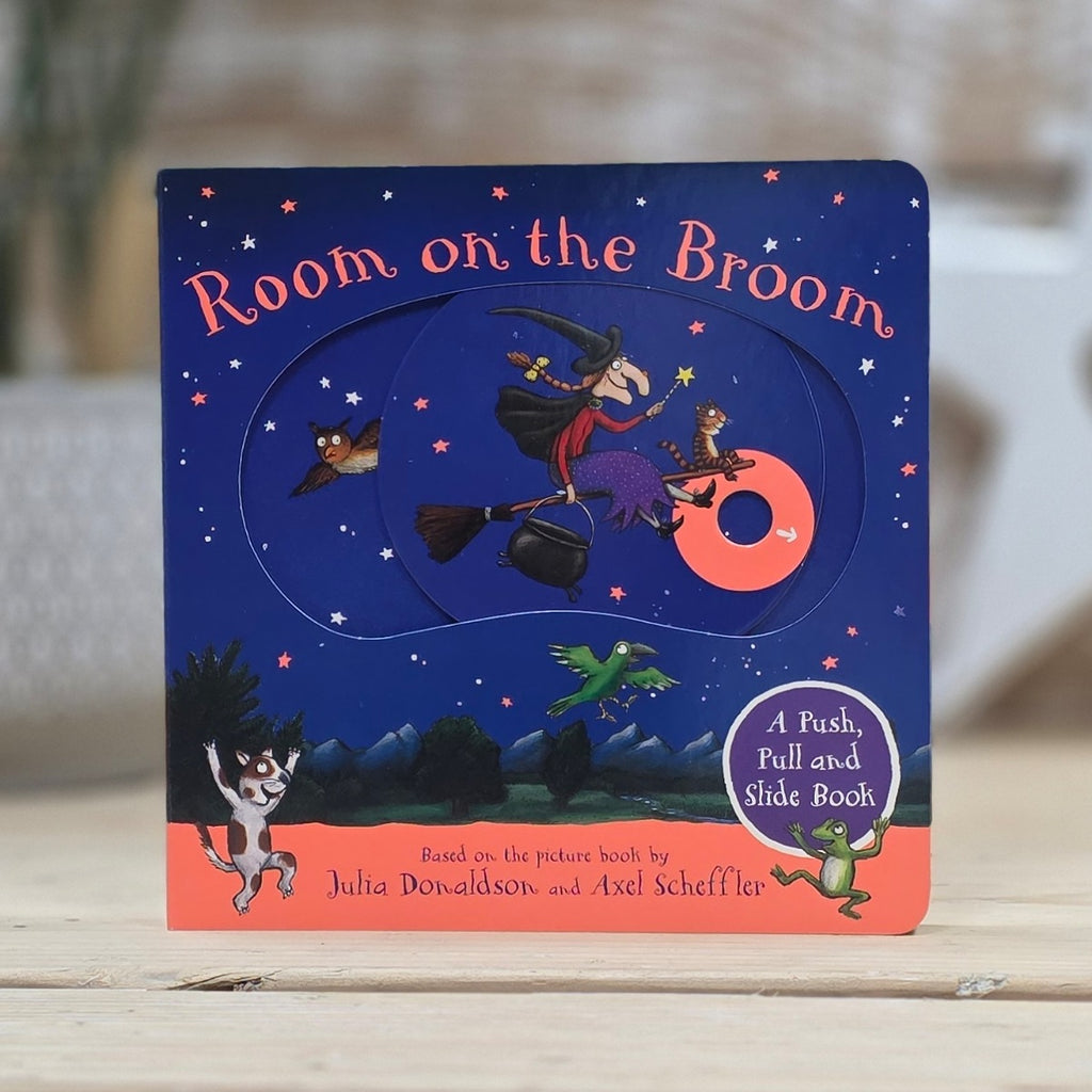A children's board book titled "Room on the Broom" standing upright on a light wood surface. The cover shows a dark blue night sky with stars, a witch riding a broom, a slide-out moon, and cartoon characters.