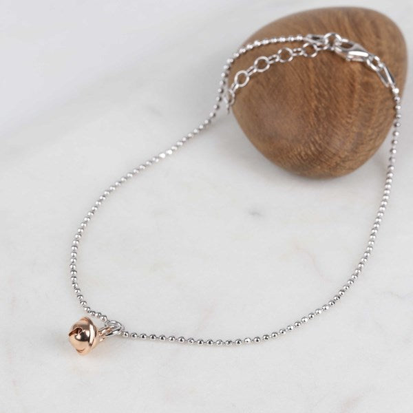 A delicate silver chain anklet with a small rose gold heart charm, displayed on a white marble surface next to a wooden block.