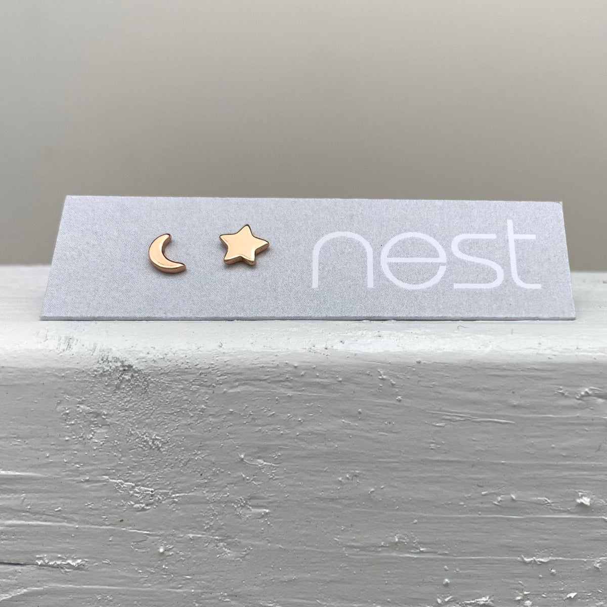 A pair of small rose gold stud earrings, one shaped like a crescent moon and the other like a five-pointed star, displayed on a light gray card with the brand name 'nest' printed on it, resting on a textured white surface.