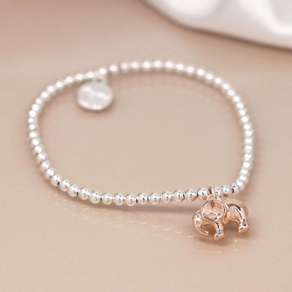 A delicate beaded bracelet featuring small silver-toned round beads, a small round silver disc charm, and a polished rose gold elephant pendant, displayed on a warm beige surface.