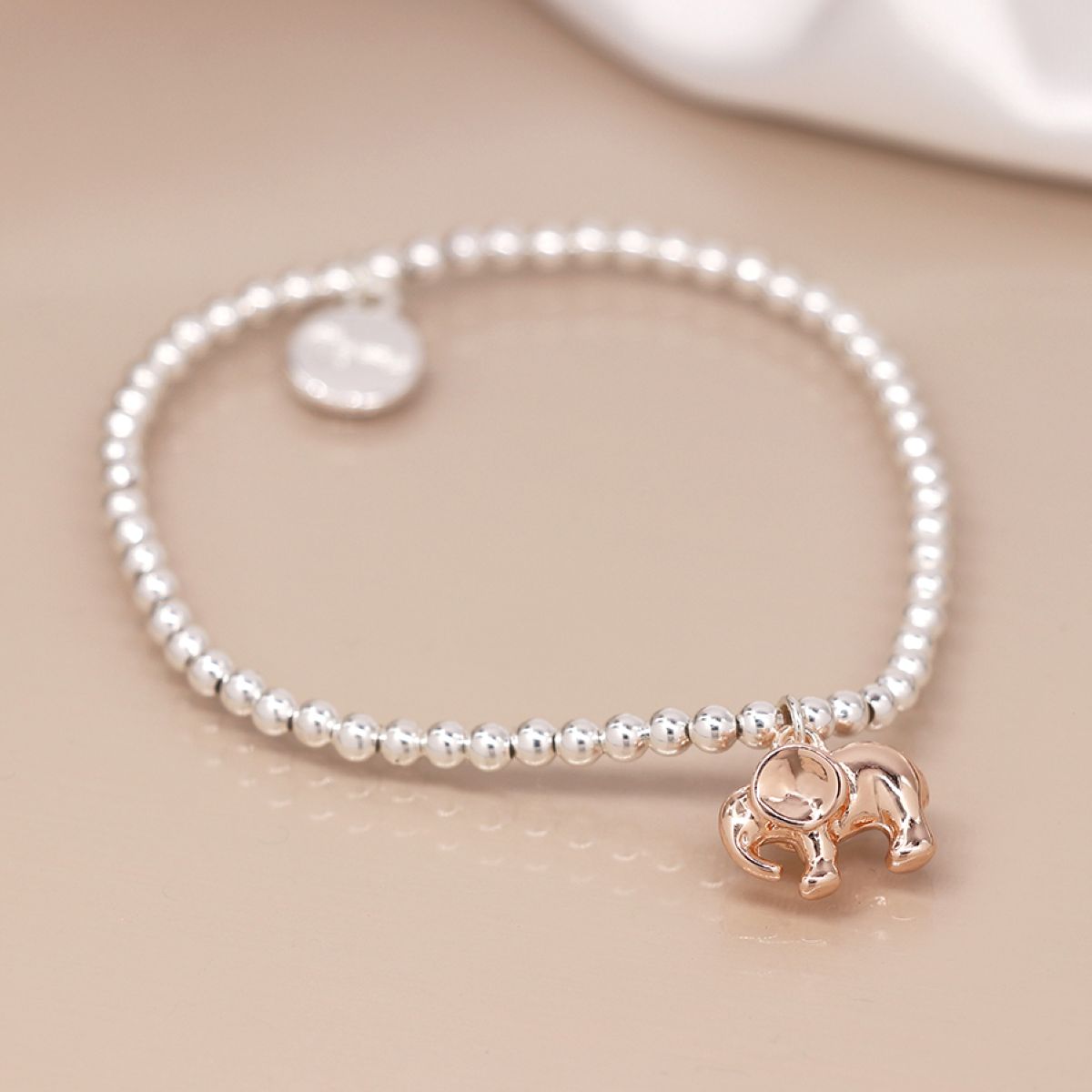 A delicate beaded bracelet featuring small silver-toned round beads, a small round silver disc charm, and a polished rose gold elephant pendant, displayed on a warm beige surface.
