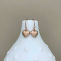 A pair of rose gold heart-shaped dangle earrings displayed on a white textured ceramic vase against a neutral beige background.