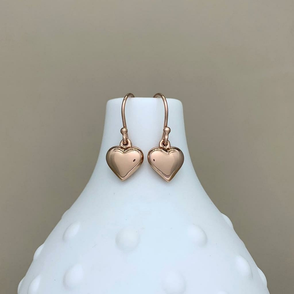 A pair of rose gold heart-shaped dangle earrings displayed on a white textured ceramic vase against a neutral beige background.