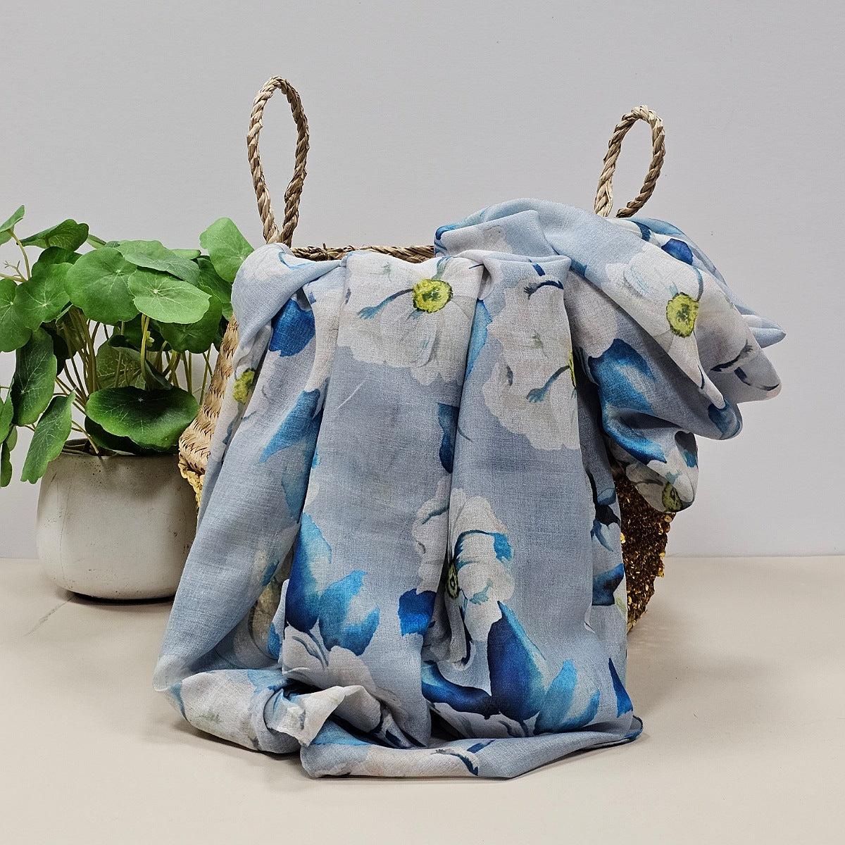 Silver scarf with floral-patterns in white, blue and yellow draped over a woven basket with a plant in the background
