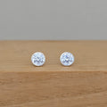 A pair of round silver stud earrings with clear faceted gemstones, resting on a light brown wooden surface against a neutral gray background.