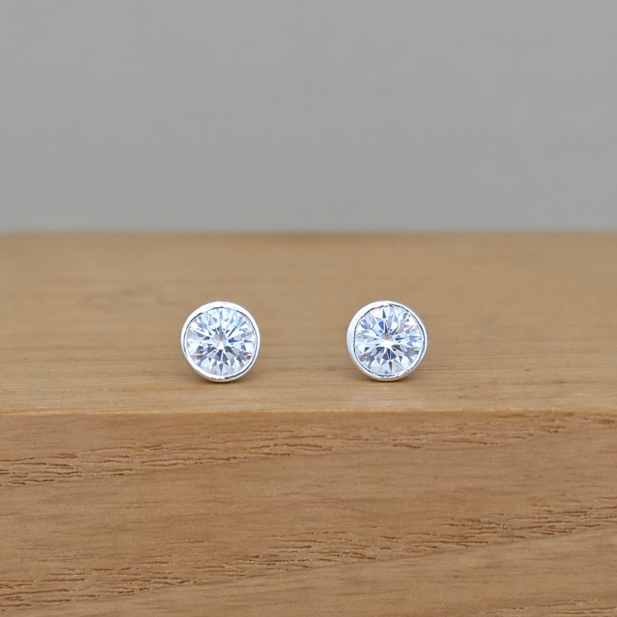 A pair of round silver stud earrings with clear faceted gemstones, resting on a light brown wooden surface against a neutral gray background.
