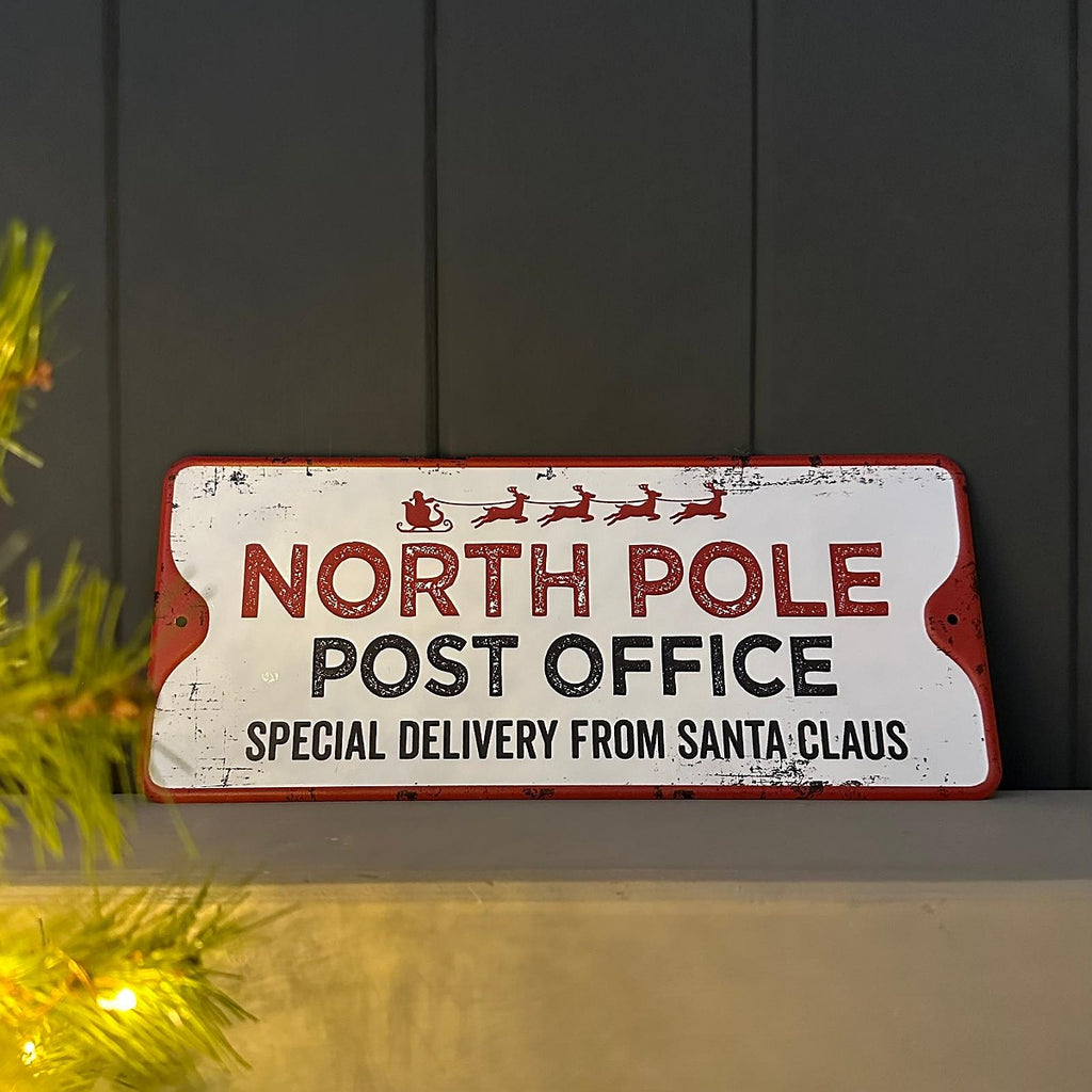 Decorative white sign stating 'North Pole Post Office, special delivery from Santa Claus' in red and black with Santa Claus sledge and reindeers  against a dark background.