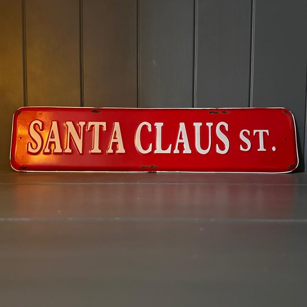 A vintage-style red metal street sign reading 'SANTA CLAUS ST.' in raised white lettering, displayed against a dark panelled wall and grey surface.