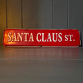 A vintage-style red metal street sign reading 'SANTA CLAUS ST.' in raised white lettering, displayed against a dark panelled wall and grey surface.