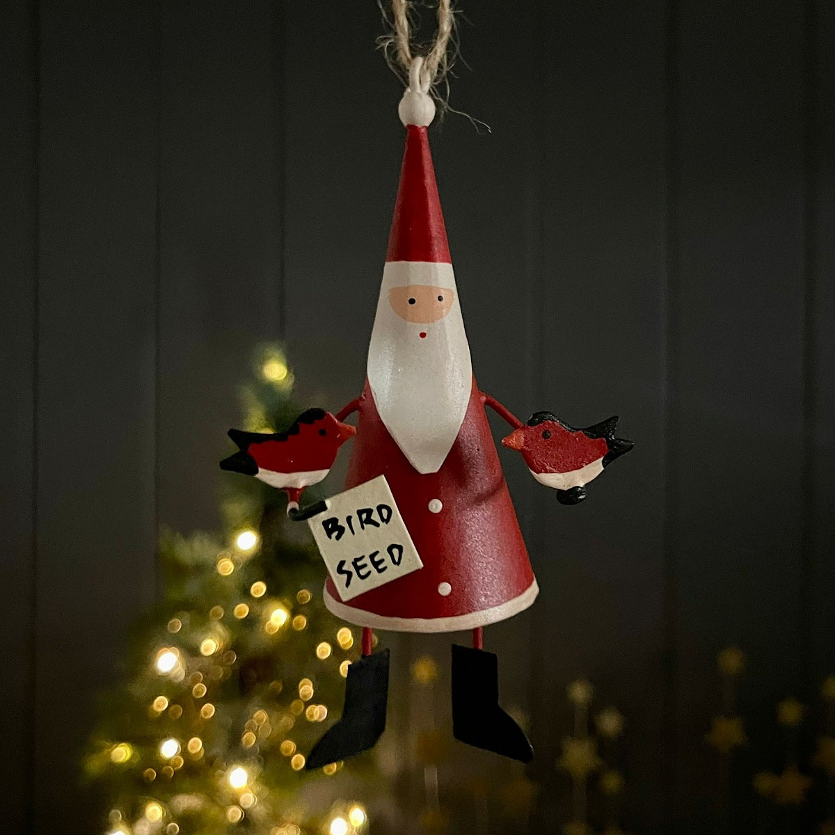A hanging metal Christmas ornament shaped like a Santa figure in a red suit with a white beard, holding two red birds and a sign that says 'BIRD SEED'. The ornament is photographed against a dark panelled background with blurred warm lights from a Christmas tree behind.