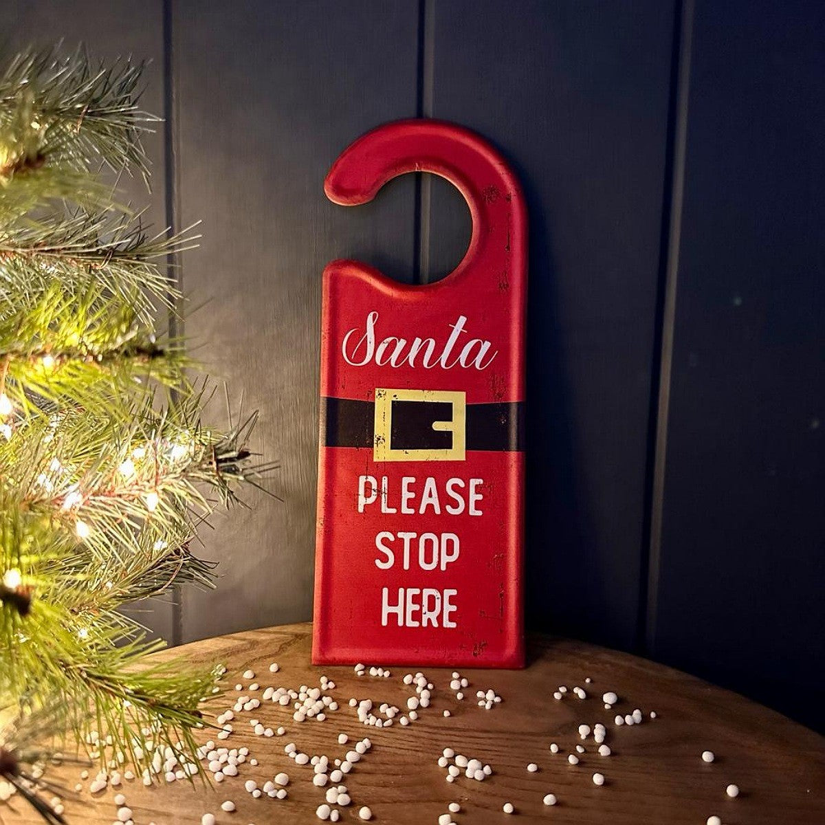 A red Christmas-themed door hanger sign with 'Santa' in white script, a black belt graphic with gold trim, and the text 'PLEASE STOP HERE' in white. The sign is displayed on a wooden surface with scattered white beads, next to a lit pine branch, against a dark panelled background.