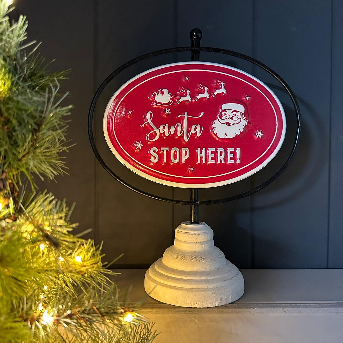 A decorative oval sign on a white pedestal base, featuring a red background with white Santa and sleigh artwork and the words 'Santa STOP HERE!'. The sign is displayed indoors against a dark paneled wall, with a lit Christmas tree branch entering from the left.