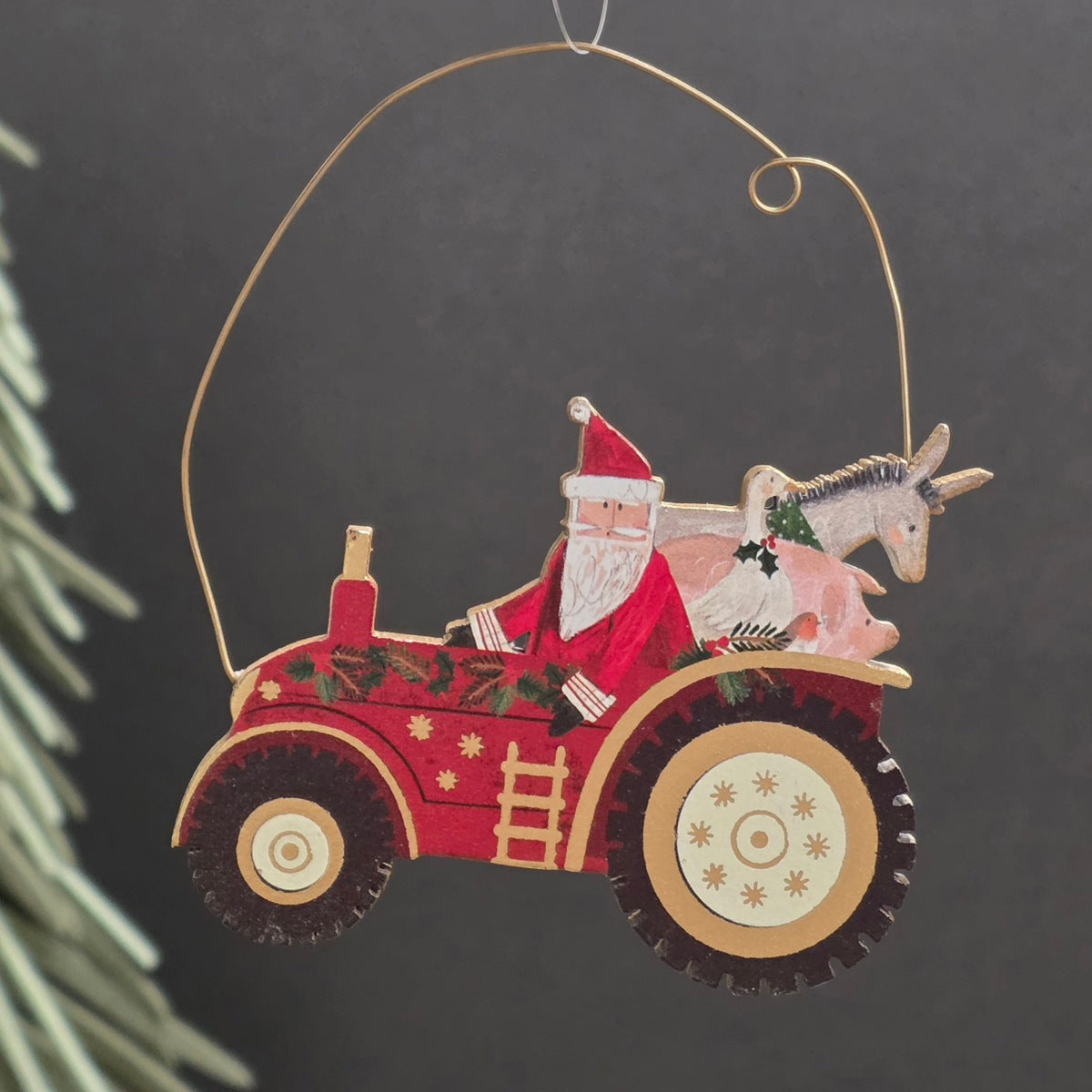 A handcrafted wooden Christmas ornament shaped like a red tractor carrying Santa Claus and a donkey and a pig, hanging from a curved gold wire against a dark grey background.