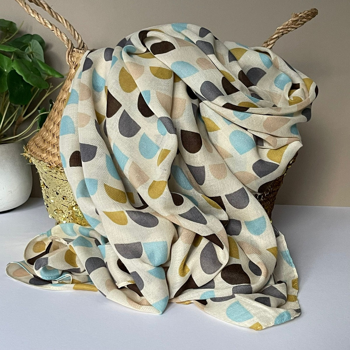 A lightweight cream scarf with a geometric semicircle pattern in mustard, light blue, taupe, grey and dark brown, draped over a woven basket next to a potted plant.