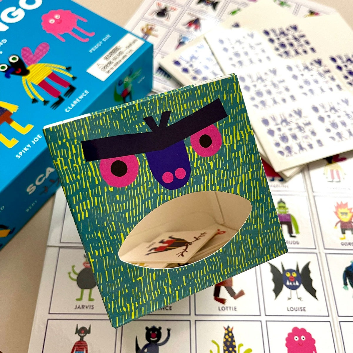 Colourful card box with a scary face and opening mouth slot with cards inside on a table with other cards and a box.