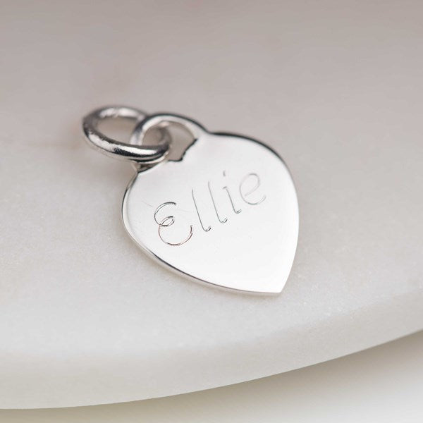 A polished silver heart-shaped charm engraved with the name 'Ellie' and attached to a matching silver jump ring, resting on a soft, light-colored surface.