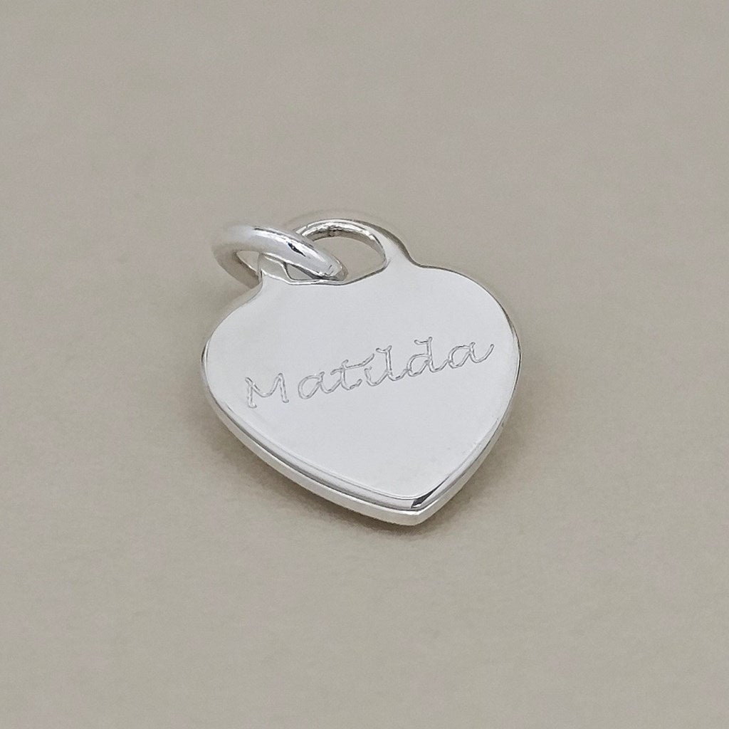 A polished silver heart-shaped pendant charm engraved with the name 'Matilda' in cursive, resting on a neutral beige background.