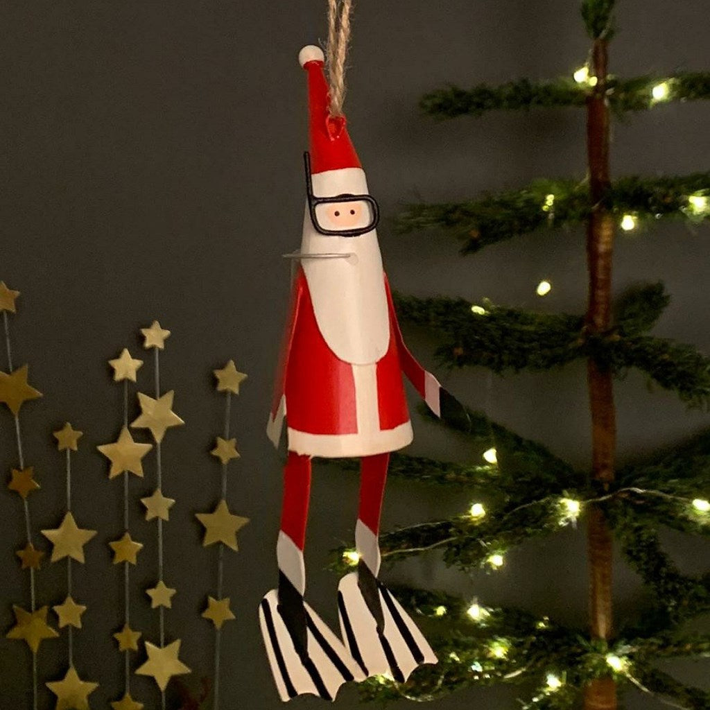 Red and white metal Santa Claus decoration with scuba diving mask, snorkel and flippers hanging on a string with a Christmas tree in the background.