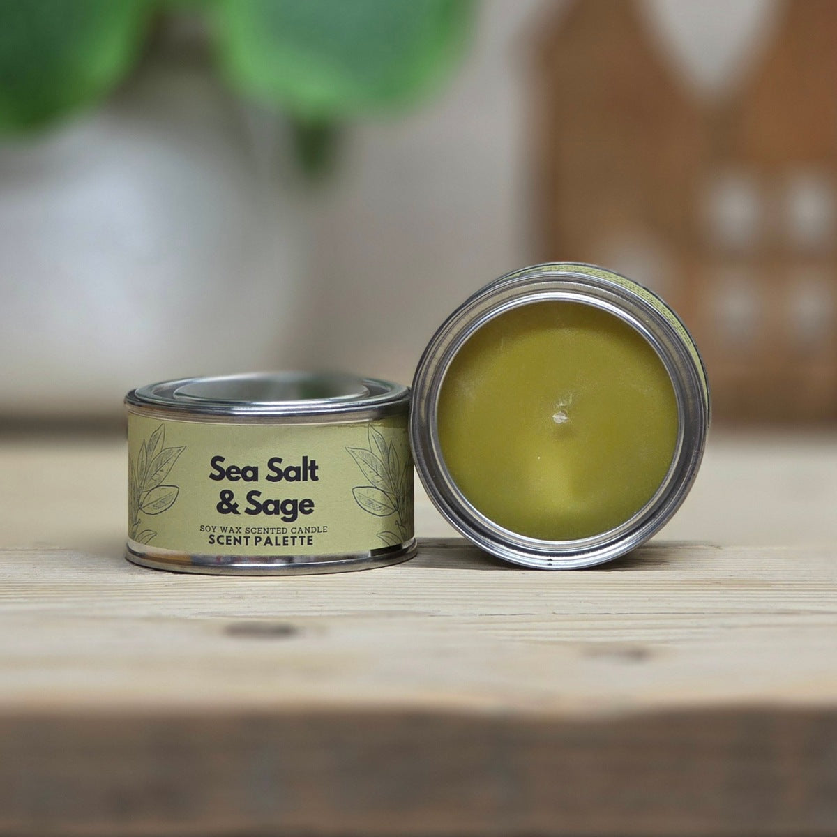 Sage green tinned candle on a wooden surface, one closed with a beige label reading 'Sea Salt & Sage' and one open showing a yellow-green wax inside.