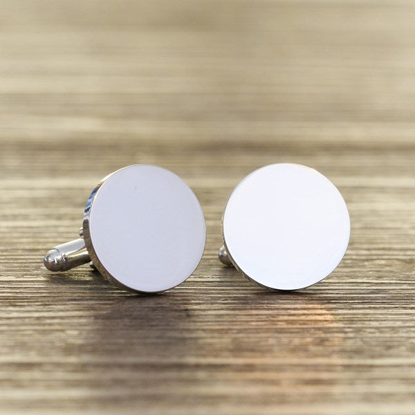 A pair of round silver cufflinks with blank silver faces, resting on a wooden surface.