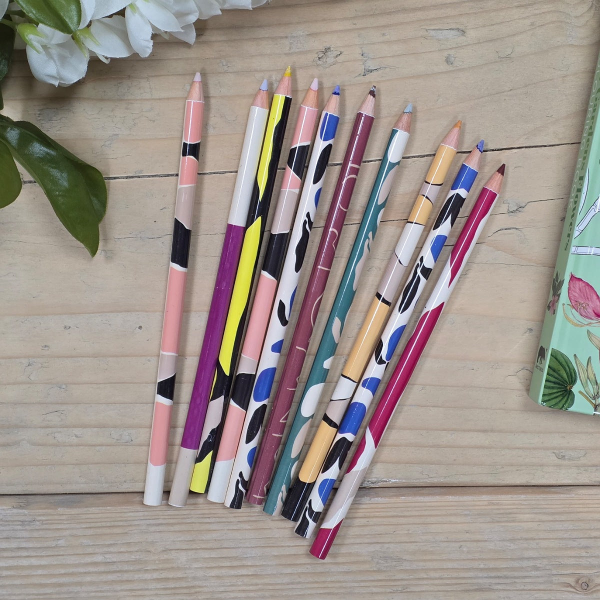 A set of ten colored pencils with colorful abstract patterns, arranged diagonally on a light wooden tabletop next to white flowers.