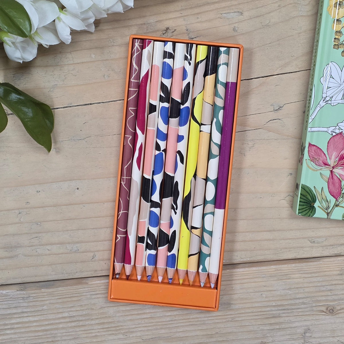 A set of 12 patterned colored pencils in an orange rectangular box, displayed on a light wood tabletop with decorative white flowers and a floral notebook nearby.