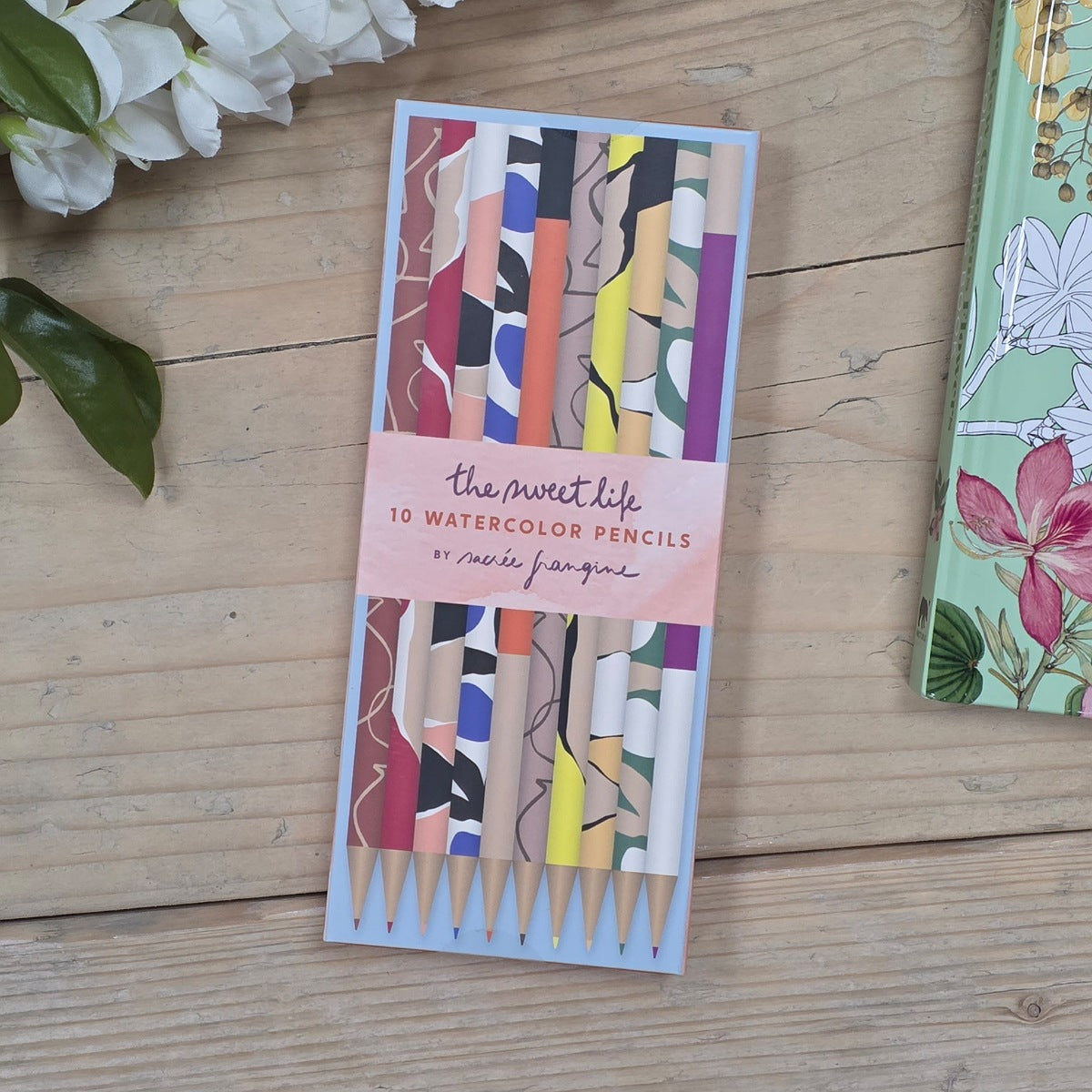 A set of 10 watercolor pencils in a light blue rectangular case, wrapped with a pink paper band reading 'The Sweet Life 10 WATERCOLOR PENCILS'. The pencils feature colorful abstract patterns.