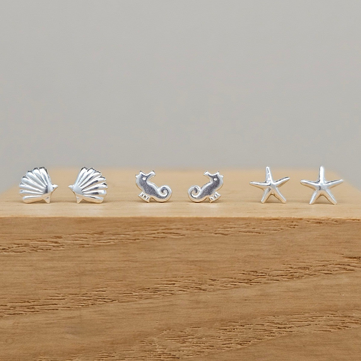 Six small silver seashell and seahorse stud earrings arranged in a row on a wooden block against a plain light gray background.