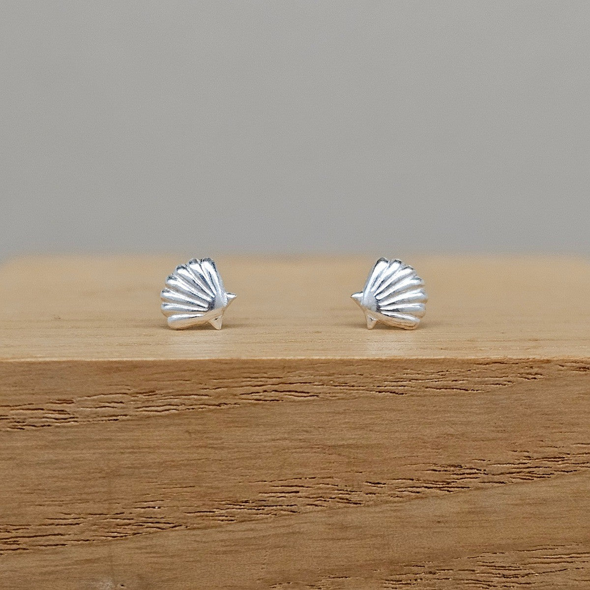 A pair of silver stud earrings shaped like small fan or shell motifs, displayed on a wooden block against a plain light gray background.