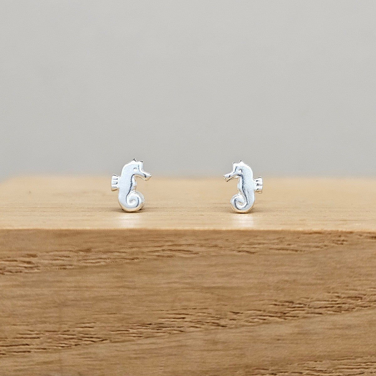 A pair of small silver seahorse stud earrings displayed on a wooden block against a neutral gray background.