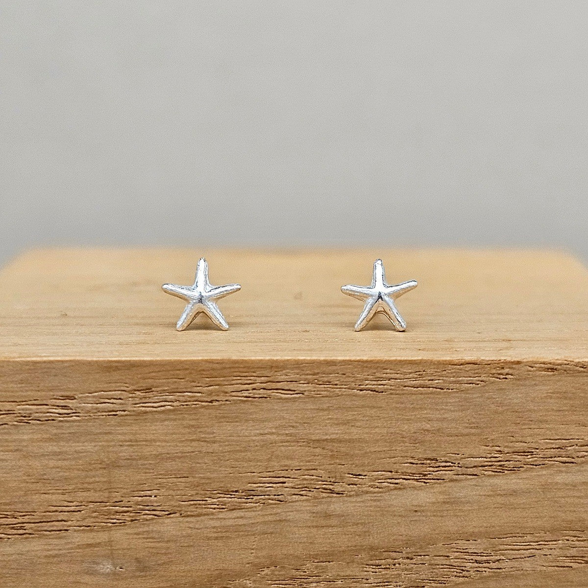 A pair of small silver starfish stud earrings resting on a wooden block against a neutral gray background.