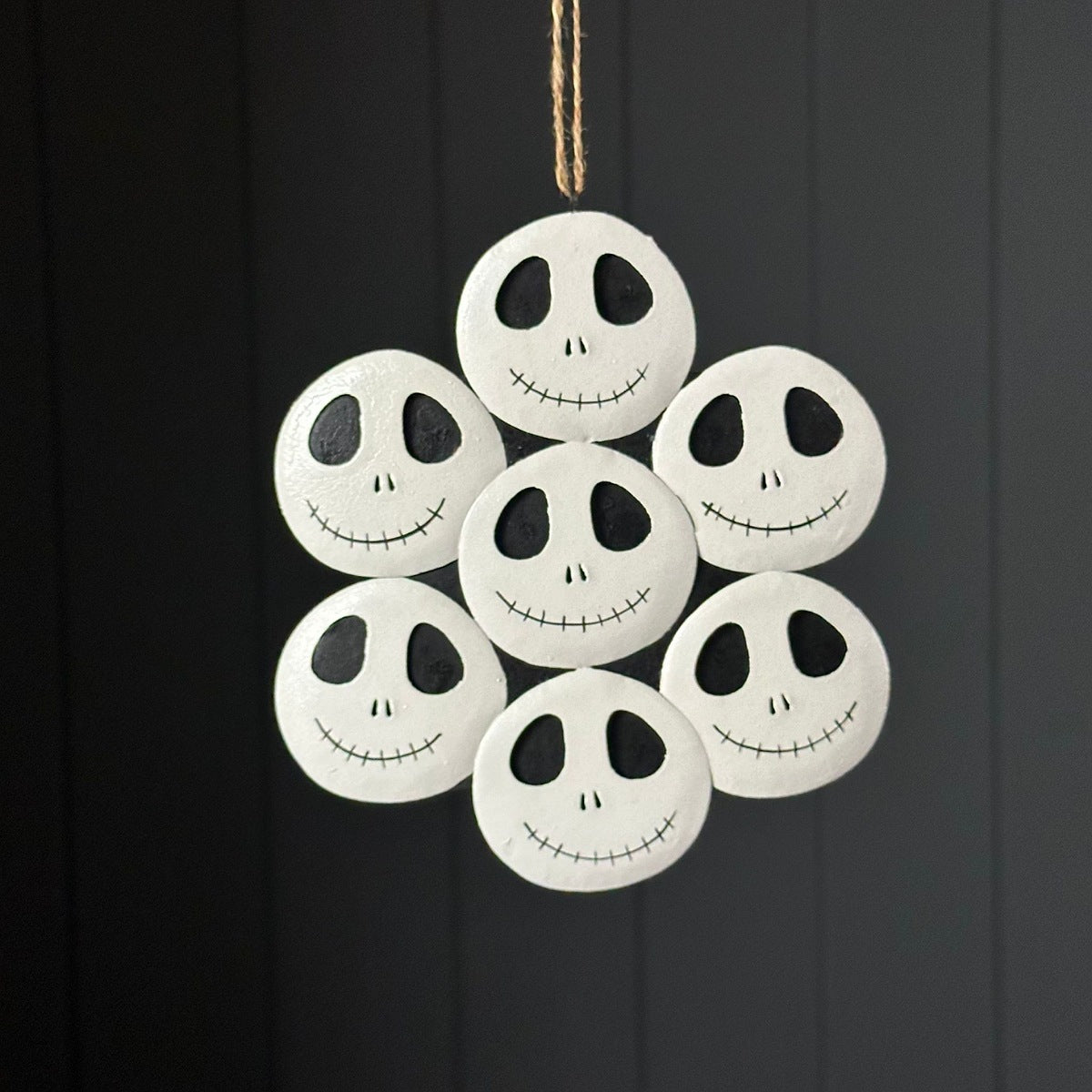 A cluster of seven white metal skeleton faces hanging with black cartoon-style faces, suspended by a thin string against a dark panelled background.