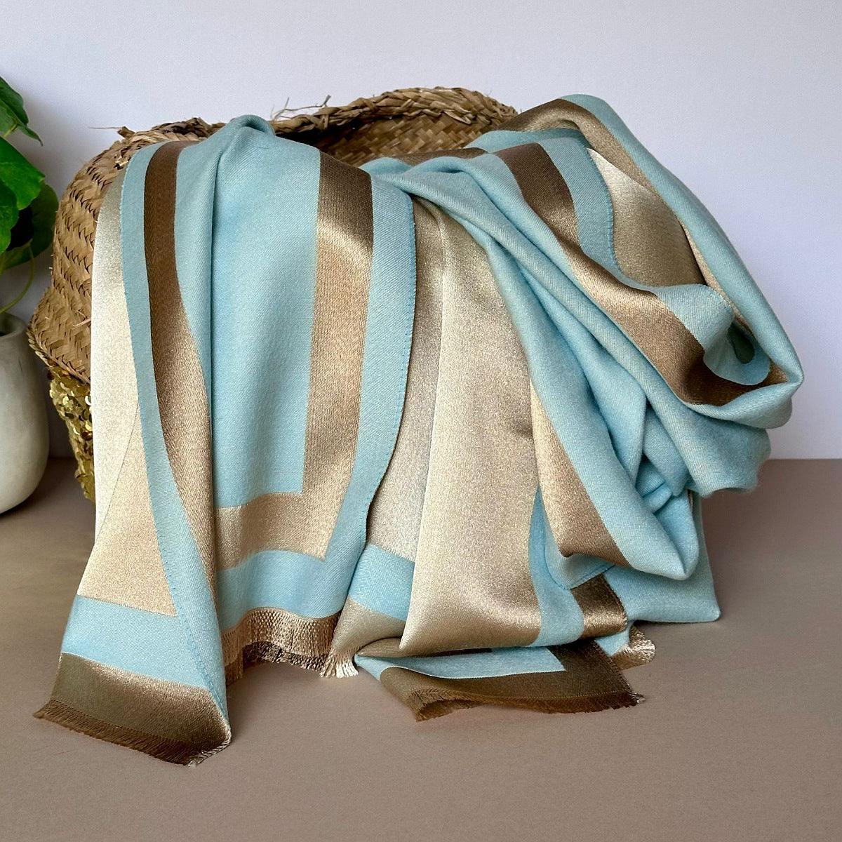 A reversible shimmering gold and light blue scarf with a contrasting boarder draped over a basket shown against a white background and potted plant to the side.