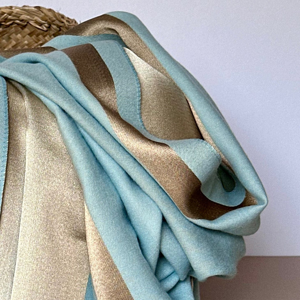 A reversible shimmering gold and dark grey scarf with a contrasting boarder draped over a basket shown against a white background.