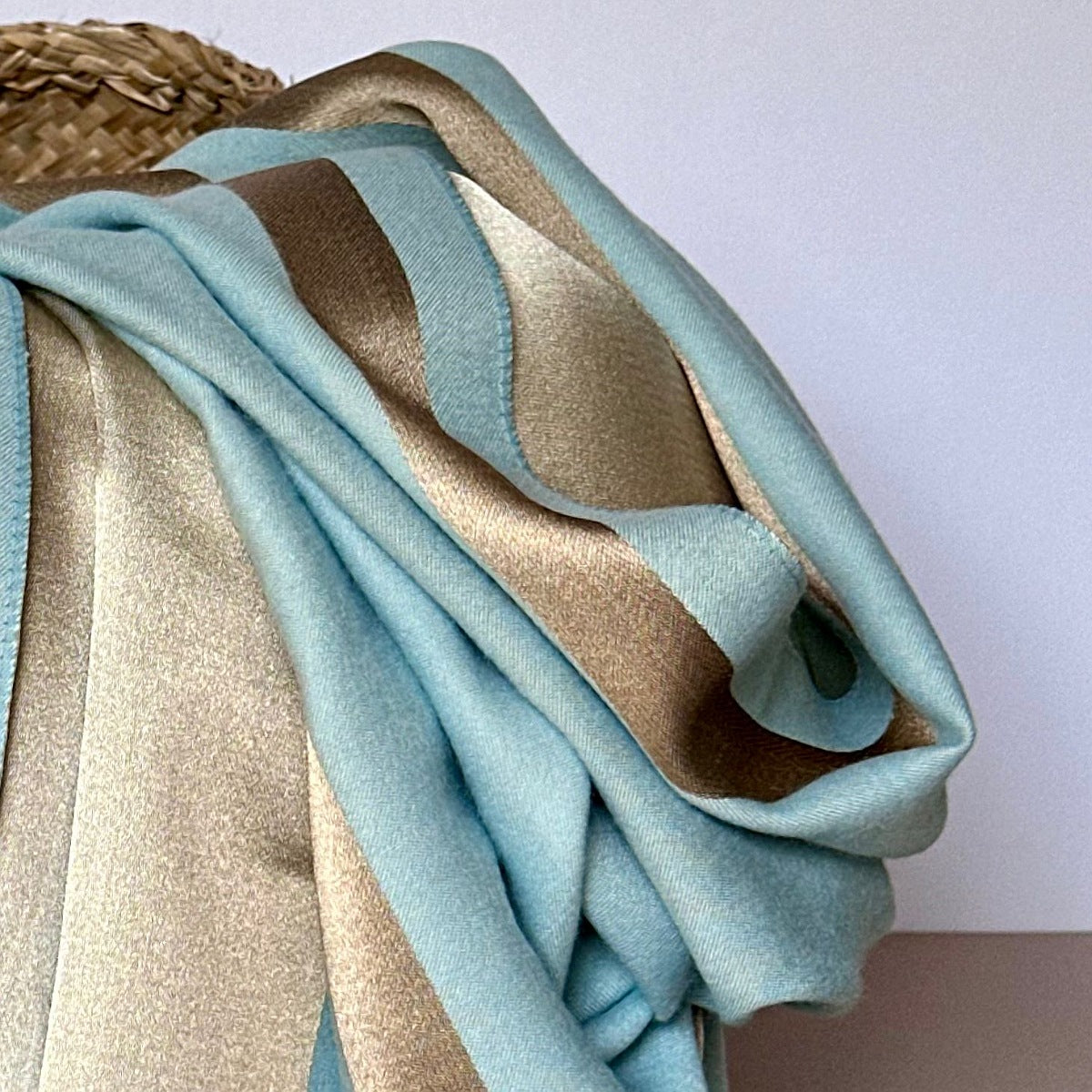 A reversible shimmering gold and dark grey scarf with a contrasting boarder draped over a basket shown against a white background.