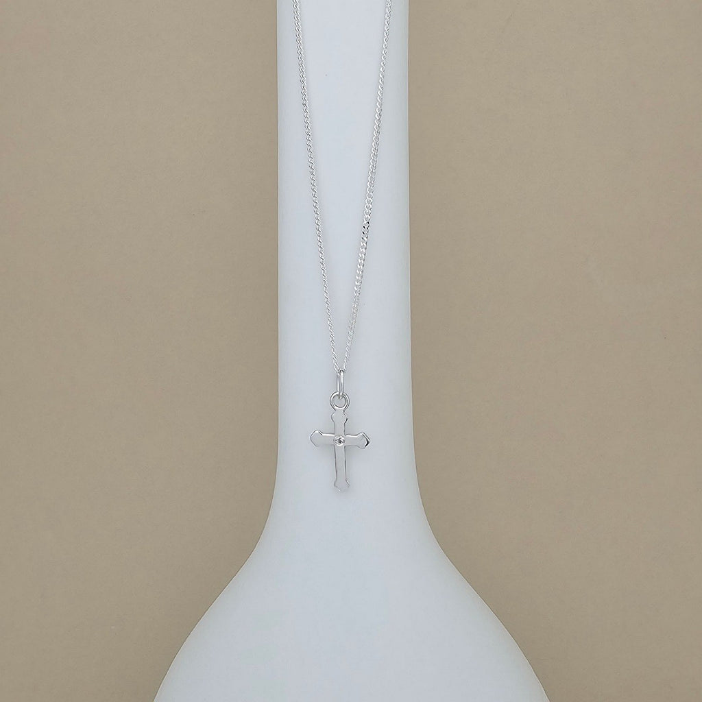 A silver cross pendant necklace hanging from a delicate silver chain, displayed on a white ceramic vase against a solid beige background.