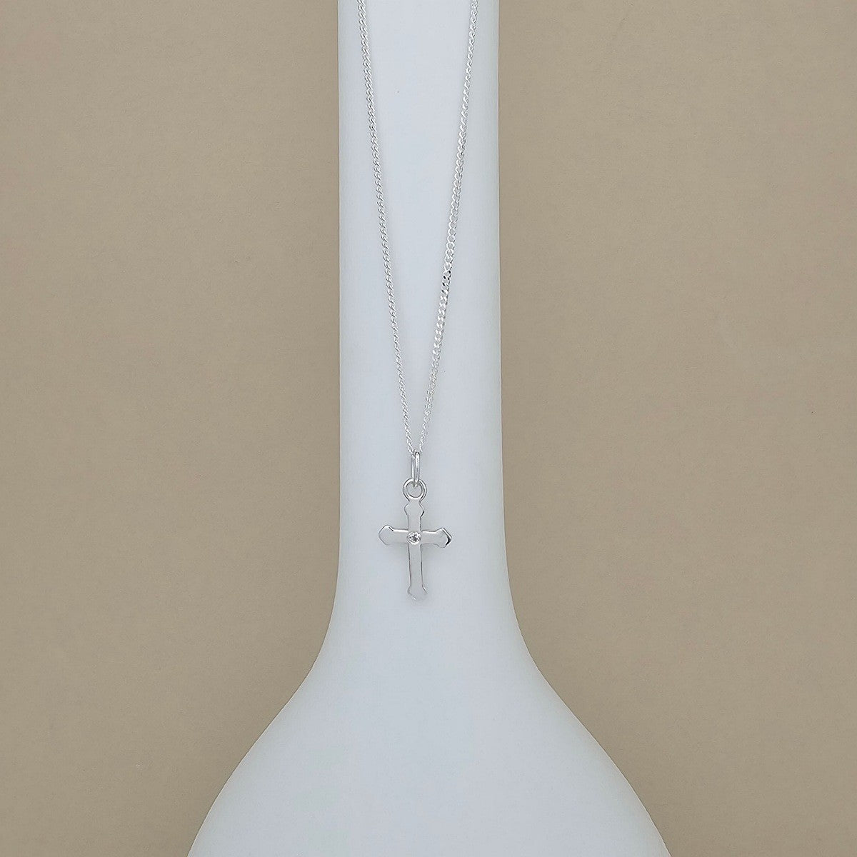 A silver cross pendant necklace hanging from a delicate silver chain, displayed on a white ceramic vase against a solid beige background.